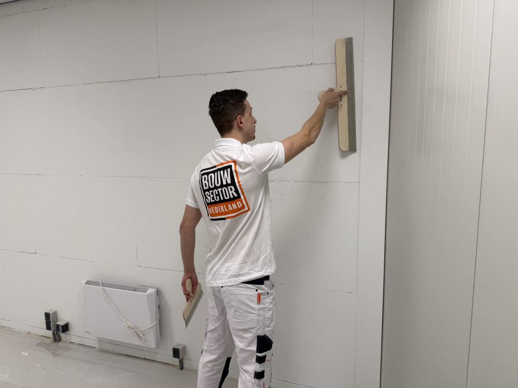 Stukadoor van Bouwsector Nederland met spaan en plamuurmes aan een lange wand bij een radiator